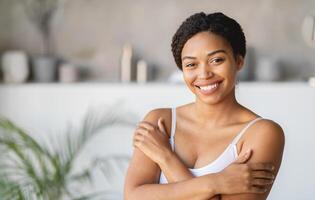A cheerful young woman stands in a well-lit modern room, wrapping her arms around herself with a warm smile. The setting features soft colors and minimalist decor that create a cozy atmosphere. photo