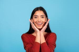 A woman with long brunette hair displays a joyful expression while resting her hands on her cheeks. She is wearing a red sweater and stands in front of a bright blue background, radiating positivity. photo