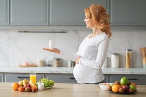 A smiling pregnant woman stands in a stylish kitchen, holding a vitamin supplement in one hand while surrounded by an array of fresh fruits and a glass of juice on the table. photo