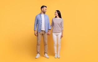 A Caucasian couple stands together against a vibrant yellow background, holding hands and smiling at each other with warmth and affection, showcasing their bond in a cheerful atmosphere. photo