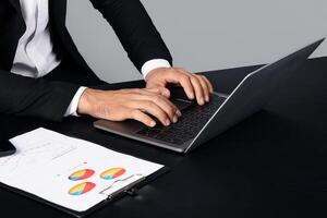 Unrecognizable man in business attire is focused on his laptop, analyzing data. Nearby, a document with colorful pie charts is neatly arranged on the table. photo