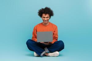 A young African American man with curly hair sits on the floor, smiling while using a laptop. He wears a casual outfit and the background is a solid light blue color. photo