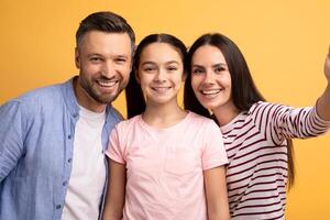 A happy family consisting of a father, mother, and teenage daughter poses together, sharing smiles and affection against a vibrant yellow backdrop. Their love is evident. photo