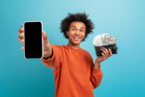 A young African American man proudly displays his smartphone in one hand and a wallet filled with cash in the other hand. He has curly hair and a bright smile, set against a blue background. photo