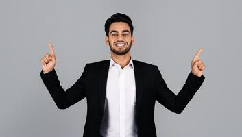 A young Arab businessman dressed in a suit stands in a studio environment, smiling and gesturing with both hands raised. His expression reflects confidence and excitement in a corporate lifestyle. photo