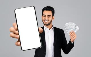 A middle eastern businessman smiles broadly while holding a smartphone in one hand and a stack of cash in the other. He is dressed professionally in a suit against a plain gray background. photo