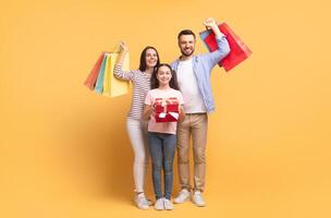 A parent and their teenage daughter joyfully pose with shopping bags in vibrant colors. They hold a red gift box, capturing a moment of love and togetherness against a bright backdrop. photo