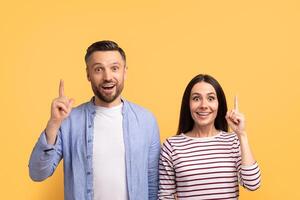 A happy couple stands together against a vibrant yellow background, both pointing upwards with joyful expressions. Their close bond and affection are evident as they celebrate a moment. photo