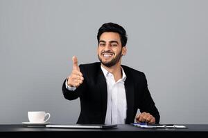 A young Middle Eastern man dressed in a suit extends his hand for a handshake, smiling warmly. He sits at a desk in a modern studio, ready for professional engagement. photo
