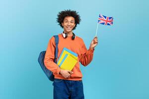 A smiling African American man holds a small British flag while carrying folders and wearing headphones. He stands against a light blue backdrop, exuding joy and enthusiasm. photo