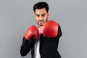 A millennial Arab entrepreneur poses confidently in a formal suit while wearing boxing gloves. He stands in a studio with a neutral background, showcasing a blend of professionalism and vigor. photo