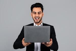 A Middle Eastern businessman smiles broadly while working on his laptop in a minimalistic studio. Dressed in formal attire, he shows excitement over a recent achievement. photo