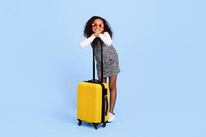 A joyful African American girl stands with a yellow suitcase, leaning on it with a big smile. She wears stylish sunglasses and is dressed in a playful outfit, exuding excitement for her adventure. photo