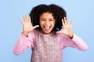 A joyful girl with curly hair displays her hands enthusiastically, expressing fun and happiness. She is dressed in a pink top and stands in front of a solid blue backdrop, radiating positivity. photo