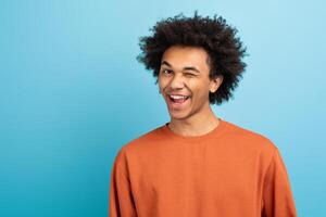 A young African American man with curly hair winks playfully while smiling. He wears an orange shirt and stands in front of a vibrant blue background, conveying a lively, fun atmosphere, copy space photo