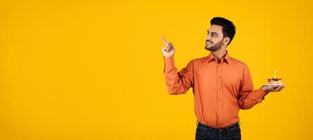 A millennial Arab man stands in a bright studio, holding a small cake with a candle. He points at copy space with a joyful expression, celebrating a personal or professional milestone. photo