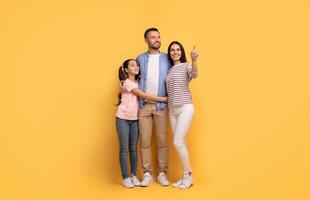 A European family stands together in front of a yellow backdrop, with the daughter hugging her father. The mother points out something, reflecting joy and connection among them. photo