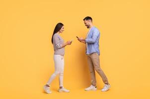 A family couple stands close together against a vibrant yellow backdrop, engaging with their smartphones. They share smiles and laughter, reflecting their affectionate bond and enjoyment. photo