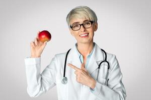 Proper diet for health is doctor recommendation. Smiling woman in white coat points finger at red apple in hands isolated on blue background, studio shot photo