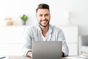 Successful freelancer. Happy young man sitting at workplace with laptop and smiling to camera while working on computer at home office, copy space. Guy enjoying remote work and self-employment photo