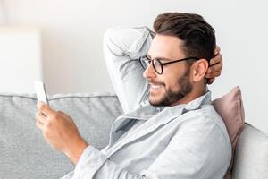 Cool gadget and app. Young smiling guy holding mobile phone, typing sms message, resting on sofa in living room, free space. Man browsing internet, surfing web, using app photo