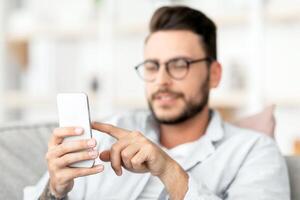 Handsome guy in eyeglasses using smatphone, touching screen while relaxing on couch at home, free space. Concentrated guy browsing internet or messaging with friends, sitting on sofa in living room photo