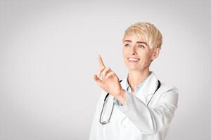 Modern medicine, diagnostics and science. Smiling adult female doctor in coat raises her hand and press fingers an empty space for virtual button, or board, or invisible screen, studio shot photo