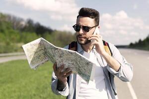 Stressed male tourist with map standing on roadside, speaking on mobile phone, cannot catch car or find his travel route. Young man feeling lost while hitchhiking on highway, having problem photo