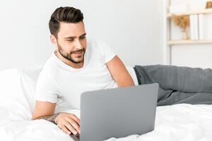 Always online. Positive young man working on laptop distantly, lying on bed, guy waking up and using pc computer, free space. Early morning and free time concept photo