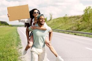 Millennial multiracial couple traveling by autostop, hitchhiking, looking for transport, trying to stop car, holding empty cardboard sign with mockup, ready strat new journey, copy space photo