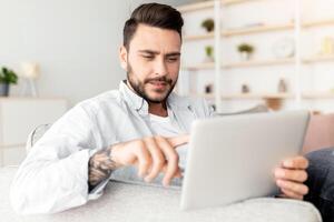 Closeup of handsome man reclining on sofa, using modern pad at home interior. Happy guy resting on couch in living room, using nice mobile app on digital tablet, copy space, panorama photo