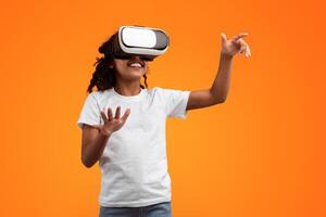 Virtual Reality Concept. Portrait of excited African American girl in VR headset touching air at yellow studio, copy space. Positive teen exploring artificial computer world or playing game photo
