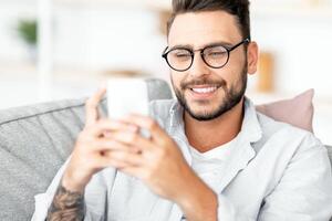 Domestic leisure. Happy guy using smartphone and resting on sofa in living room interior, smiling while messaging on cellphone, sitting on comfortable couch, copy space photo