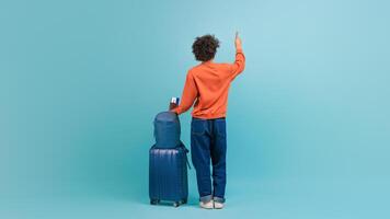A young African American man dressed in an orange sweater and jeans stands with his back to the viewer, waving goodbye. He holds a phone and has a suitcase beside him, ready for his journey. photo
