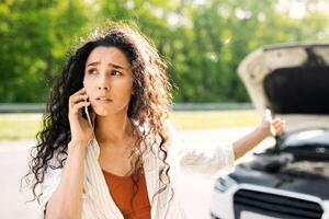 Road accident, car wreck, engine failure concept. Young woman with cellphone calling towing service on road, having phone conversation with mechanic near her broken auto on highway photo