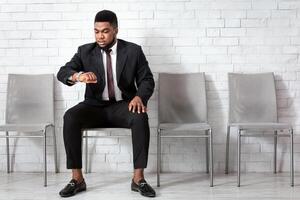 Young African American man in formal wear looking at his watch, checking time, waiting for job interview at office lobby, copy space. Personnel recruitment concept photo