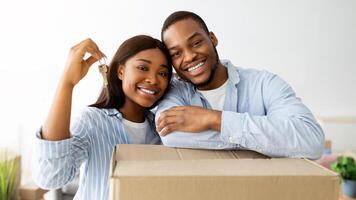 Real estate concept. Loving african american young spouses posing with moving boxes and new house key, hugging and smiling to camera, standing in room full of boxes photo