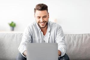 Recruitment concept. Happy millennial guy browsing work opportunities online, using job search app or website on laptop, sitting on sofa at home, free space photo