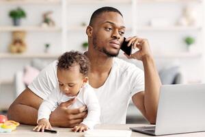 Busy Black Man Working Remotely At Home With Little Baby On Hands, African American Dad Sitting At Desk With Laptop And Talking On Cellphone While Babysitting His Infant Child, Copy Space photo