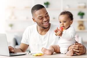 Young Black Daddy Working Online On Laptop And Playing With His Little Baby At Home, Happy African American Man Spending Time With Infant Child And Using Computer For Remote Job, Copy Space photo