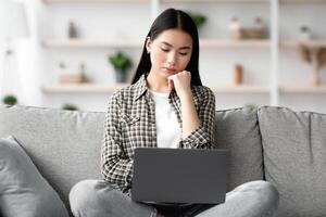 Pensive young asian lady sitting on couch and using laptop, looking for job online from home, copy space. Chinese woman student studying, reading articles on Internet, using notebook photo