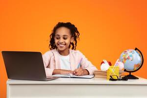 Remote Learning. Portrait of positive African American schoolgirl sitting at desk, writing in notebook and using laptop computer. Smiling young black girl watchin online course or having virtual class photo