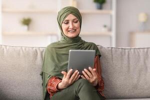 Happy Middle-Aged Muslim Lady In Hijab Using Tablet Computer Watching Movie Online Sitting On Sofa At Home. Modern Middle-Eastern Female Browsing Internet, Using Digital Gadget Indoor photo