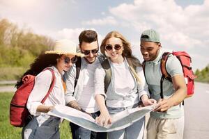 Group of best friends with backpacks checking map during road trip, hitchhiking on highway. Multiracial young tourists plotting their route, having autostop journey on summer holidays photo