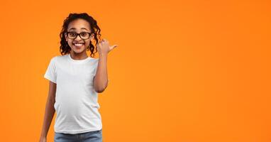 Great Offer. Portrait Of Happy Smiling African American Girl In Glasses Pointing Thumb Aside At Free Blank Space For Text Or Advert Standing Isolated On Yellow Studio Background. Look There, Banner photo