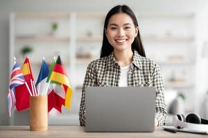 Positive asian young woman foreign language teacher sitting at workdesk with international flags and using laptop, smiling at camera, copy space. Foreign language online course, class concept photo
