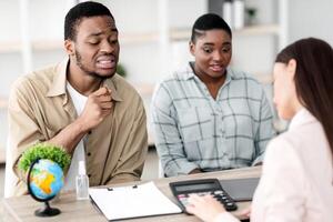 Confused Black Tourists Couple Looking At Price Of Expensive Family Tour Offered By Travel Agent Lady Sitting In Modern Office. Vacation Expenses, Financial Problems Concept. Selective Focus photo