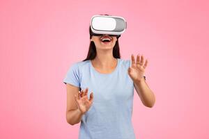 Excited young woman in VR headset touching air during virtual reality experience at studio. Emotional lady exploring artificial computer world or playing game, isolated on yellow background photo