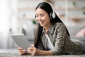 Pretty asian lady watching movie, using wireless headset and pad while laying on floor at home, closeup, copy space. Happy young woman enjoying new entertaining mobile application on digital tablet photo