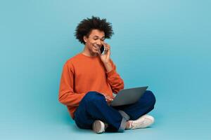 A young African American man sits cross-legged on the floor, smiling as he talks on the phone. He is using a laptop in front of him against a bright blue background. photo